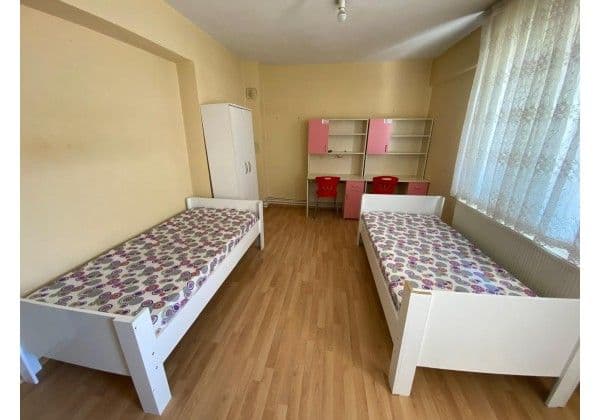 İki kişilik odada iki yatak, gardırop, kitaplık ve tül perdeli pencere